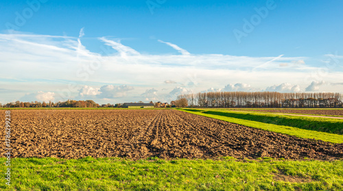 Newly ploughed field. Around it is a field margin covered with grass. In the background is a farmhouse with barns. The photo was taken at the end of a sunny autumn day.