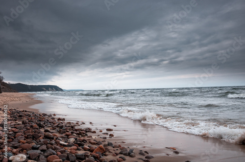 A moody coastal scene featuring a pebbled shoreline, crashing waves, and a dramatic stormy sky. The dark clouds mirror the turbulent sea, creating a sense of solitude and natural power
