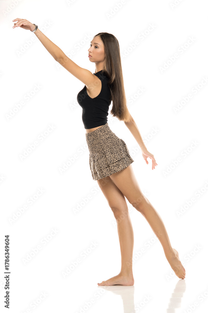 Fototapeta premium Studio shot of tall young ballerina with long brown hair in black top and leopard mini skirt posing with extended arm standing full foot on white background.