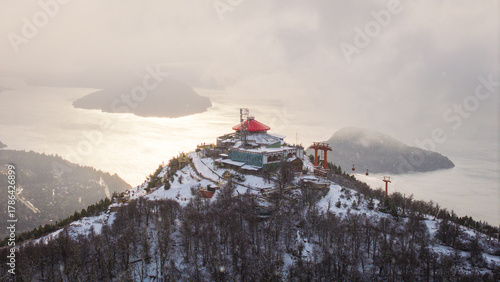 Aerial winter view of Cerro Otto with red mountain refuge and Nahuel Huapi Lake reflecting sunlight in Bariloche, Argentina.