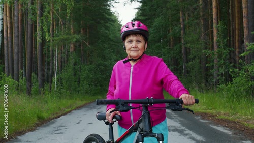 Cheerful elderly woman wearing a pink helmet and jacket smiling while standing with her bicycle on a paved road in the middle of a beautiful pine forest, enjoying an active outdoor lifestyle