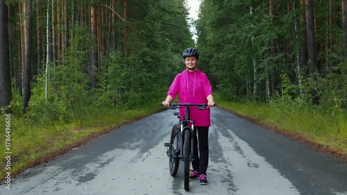 Active mature woman in a helmet walking with her mountain bike along a wet asphalt road through a beautiful green forest, enjoying a healthy outdoor lifestyle and looking at the camera