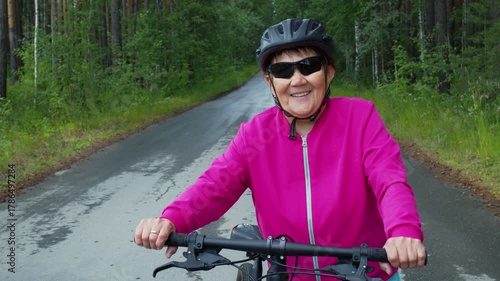 Positive elderly woman in a safety helmet and sunglasses smiling while cycling on an asphalt road through a beautiful green forest, enjoying her healthy and active retirement lifestyle