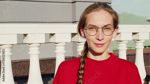 Close-up portrait of a lovely young woman with a long braid and stylish glasses, smiling sincerely and laughing happily while standing outside on a sunny day against a classic balustrade