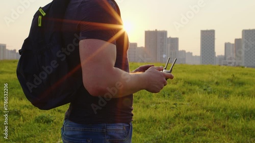 Unrecognizable drone pilot operating a remote controller in a grassy field with a modern cityscape in the background during a beautiful golden hour sunset, showing a blend of nature and technology