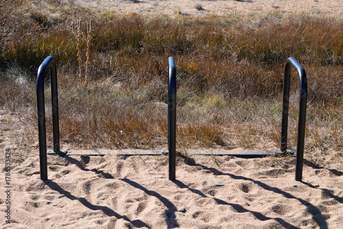Simple black metal bike rack with three U-shaped loops stands securely in the sand on a sunny beach, surrounded by sparse coastal vegetation and a clear horizon. Perfect for themes of outdoor rec.
