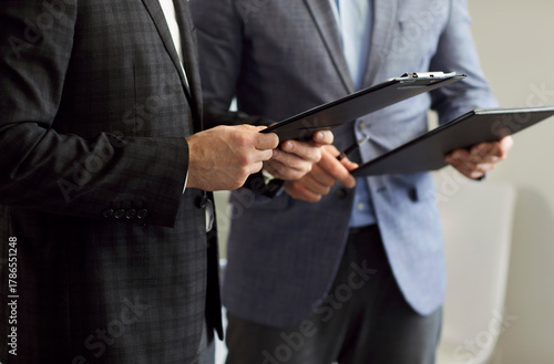 Fotografija Cropped close-up photo of two businessman in suits holding clipboards, reviewing contract details, discussing agreement terms and conditions before signing it