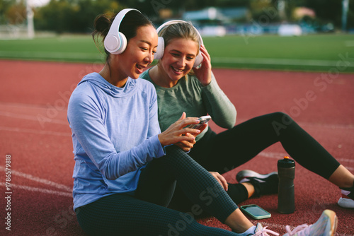 Two diverse female friends and athletes, one Asian and one Caucasian, happily share music and look at a mobile phone during a relaxed break on a running track