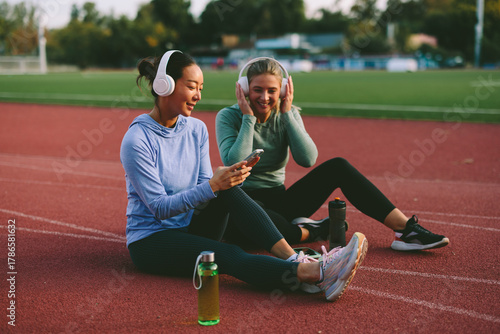 Two diverse female friends and athletes, one Asian and one Caucasian, happily listen to music on shared headphones and look at a mobile phone during a fitness break on a running track