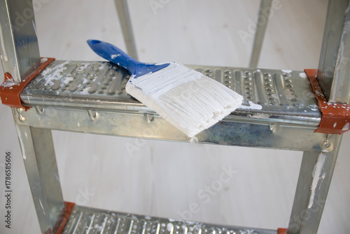 Canvas Print Detailed view of a white paint brush on a metal ladder step during indoor repainting and apartment upgrade