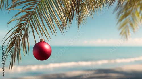 Fototapeta Naklejka Na Ścianę i Meble -  Single red Christmas ornament hanging from palm tree branch. Tropical beach panorama background