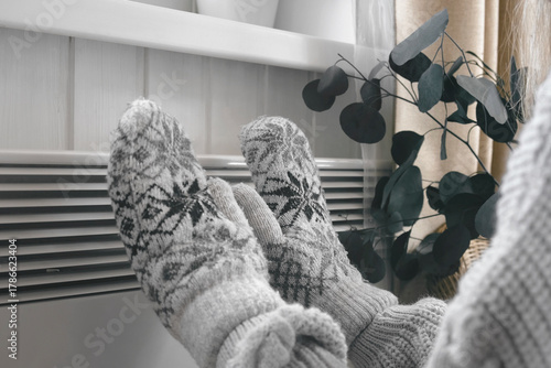 Hands in winter gloves warming up over electric heater. Frozen woman wearing a knitted mittens freezing for winter cold. Discomfort spending time at home. Girl warming hands on modern radiator.