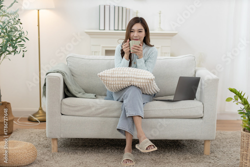 Free Download Asian women sitting on sofa at home with hot drink, holding pillow and relaxing in cozy living room. Peaceful morning moment, casual lifestyle, remote work or weekend leisure. - GFXLoad