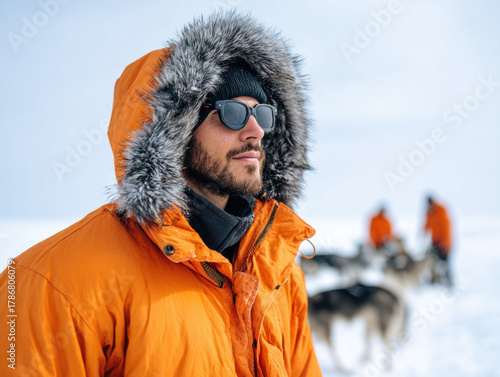 Resilient arctic explorer wearing orange parka with fur lined hood and sunglasses, surrounded by snowy landscape and sled dogs