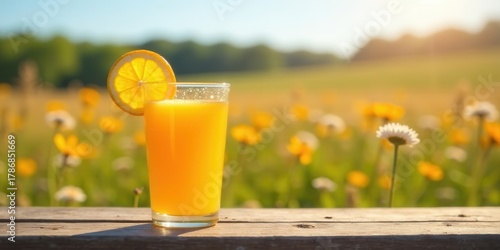 Fototapeta Naklejka Na Ścianę i Meble -  Refreshing citrus beverage in a glass, garnished with a citrus fruit slice, rests on rustic wooden surface against a vibrant sunlit meadow of wildflowers