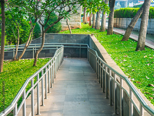 Accessibility ramp with stainless steel handrails in a public park, surrounded by green grass and trees, promoting inclusive and barrier-free urban design.