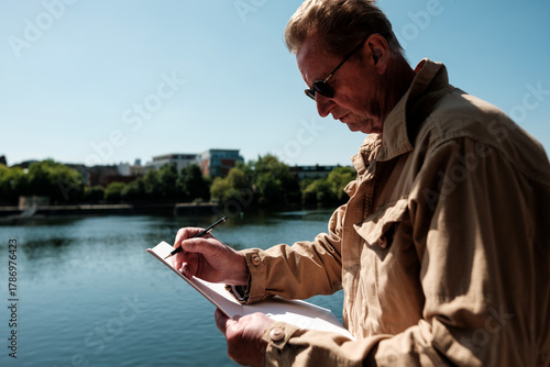 Senior engineer writing notes on clipboard near river in city