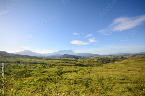 A wide, sunlit vista of expansive green grasslands and hills with Mount Kinabalu visible in the hazy distant mountains.