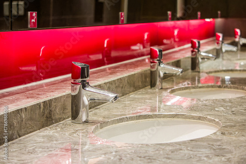 Modern Public Bathroom Sinks with Red Accent and Stone Countertop. Closeup on row of polished chrome faucets and white sinks set into luxurious marble countertop with red backsplash in public restroom