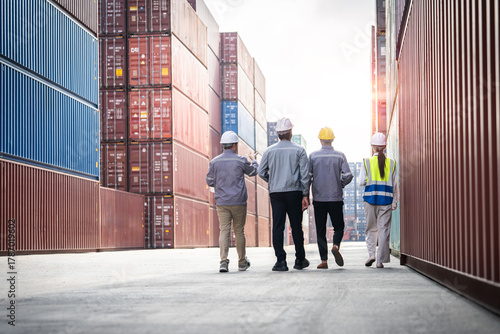 Four men in hard hats walk down a long hallway in a warehouse. They are wearing reflective vests and are likely workers in a shipping or logistics company