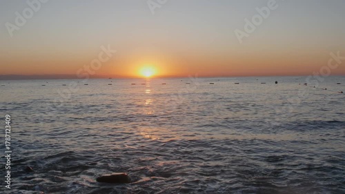 A wide, cinematic view of a quiet seascape during either sunrise or sunset. The sky is painted in soft orange and peach tones, complementing the deep blue and gray hues of the water.

