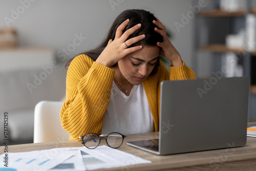 An Arab woman is sitting at her desk at home, working on a laptop. She appears stressed, touching her head, struggling with problems related to her job.