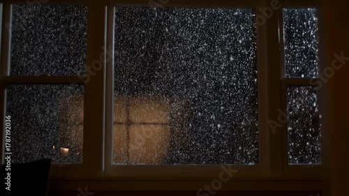 View through a window of heavy falling snow from a dark cozy room with warm indoor light reflection, seamless loop, concept of peaceful hypnotic winter isolation