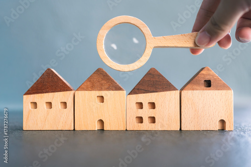 Hand holding magnifying glass over wooden houses symbolizing real estate search, property inspection, home selection, housing investment, or mortgage decision-making process.