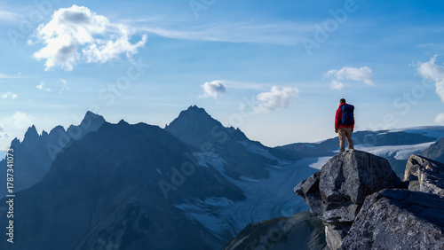 Hiker Stands on a Cliff Overlooking Jagged Mountain Range Under Blue Sky During Sunrise Adventure