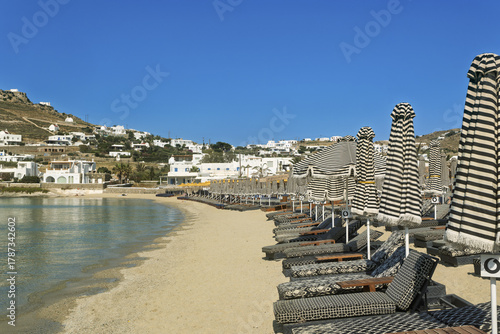 Fototapeta Naklejka Na Ścianę i Meble -  Serene beach scene in Mykonos, Greece,  with a row of black and white striped lounge chairs and matching umbrellas arranged neatly on sandy shore,