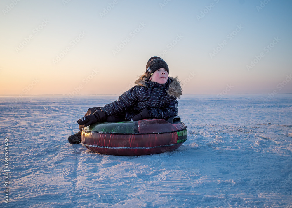 Obraz premium young boy in winter attire rests on an inflatable snow tube in a vast, snow-covered landscape under a soft, pastel-colored sky.