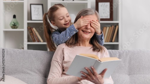 Granddaughter cheerfully covers the eyes of her grandmother who is reading a book sitting on the sofa, they laugh, play, spend time together at home.