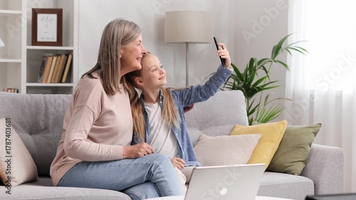 Grandmother senior lady and blonde little granddaughter taking selfie on phone sitting on sofa, looking at photos hugging.