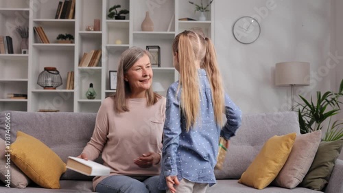 A smiling happy senior woman rejoices at an unexpected surprise gift from her little granddaughter. Grandma is reading a book without realizing that a gift is waiting for her.