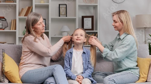 A senior woman, a grandmother, and a young mother make braids for a little blonde granddaughter on the sofa in the living room. Time spent with family, mother's day, three generations.
