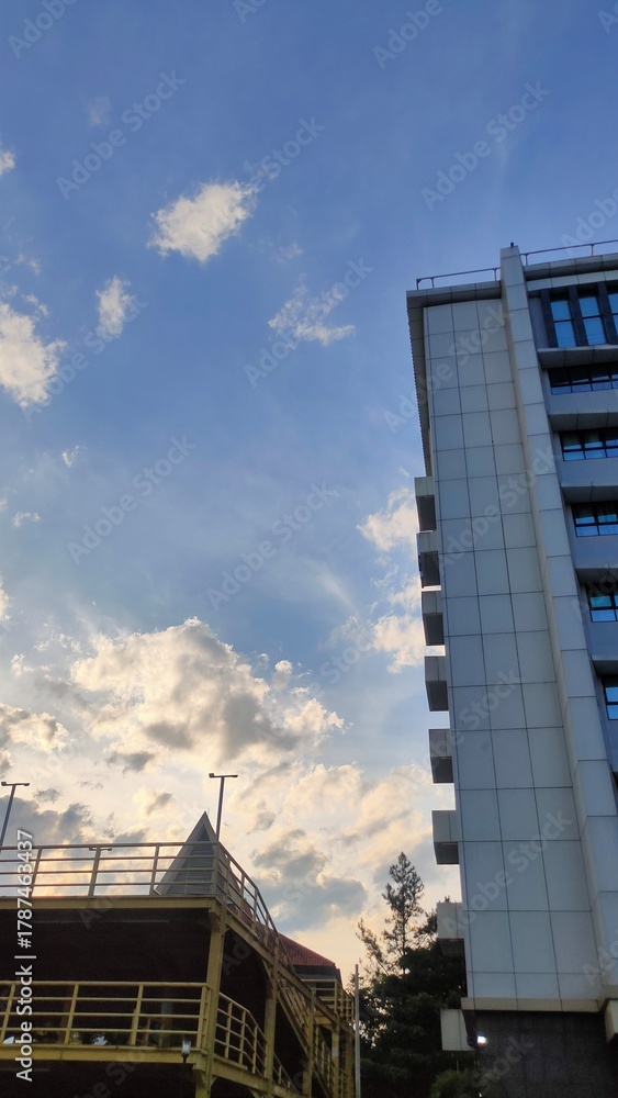 Obraz premium A tall white building stands against a bright blue sky filled with soft clouds. The warm light of the afternoon sun creates a peaceful and calming atmosphere.