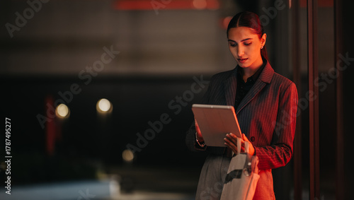 Schilderij op canvas A poised professional woman stands outdoors at night, examining content on a tablet