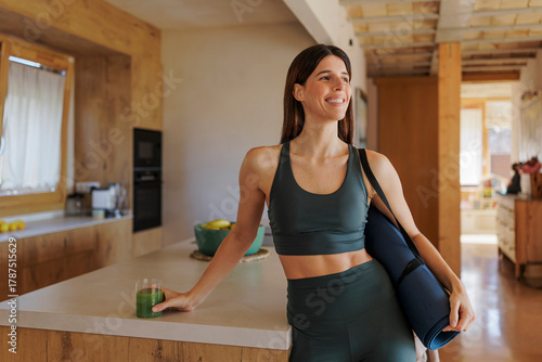 Woman in activewear with yoga mat and green juice in a modern kitchen