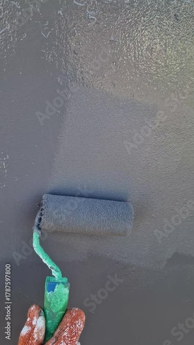 Vertical, top-down view of a gloved hand using a roller to apply dark grey paint onto a textured surface, creating a wet, uniform finish. A protective orange and white glove is visible.