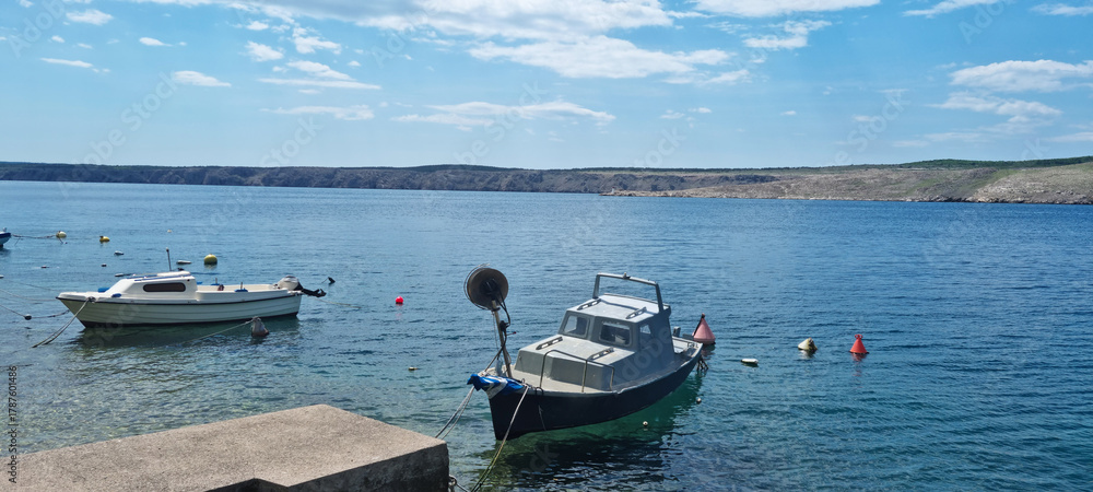 Obraz premium Two small boats are moored in the clear blue Adriatic sea off the coast of Jadranovo, with the rugged, uninhabited shore of Krk Island visible in the distance