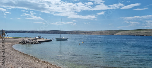 Fototapeta Naklejka Na Ścianę i Meble -  A sailboat is moored in the clear blue water of Jadranovo beach with a pebbly shore, facing the rugged coastline of Krk Island under a cloudy sky