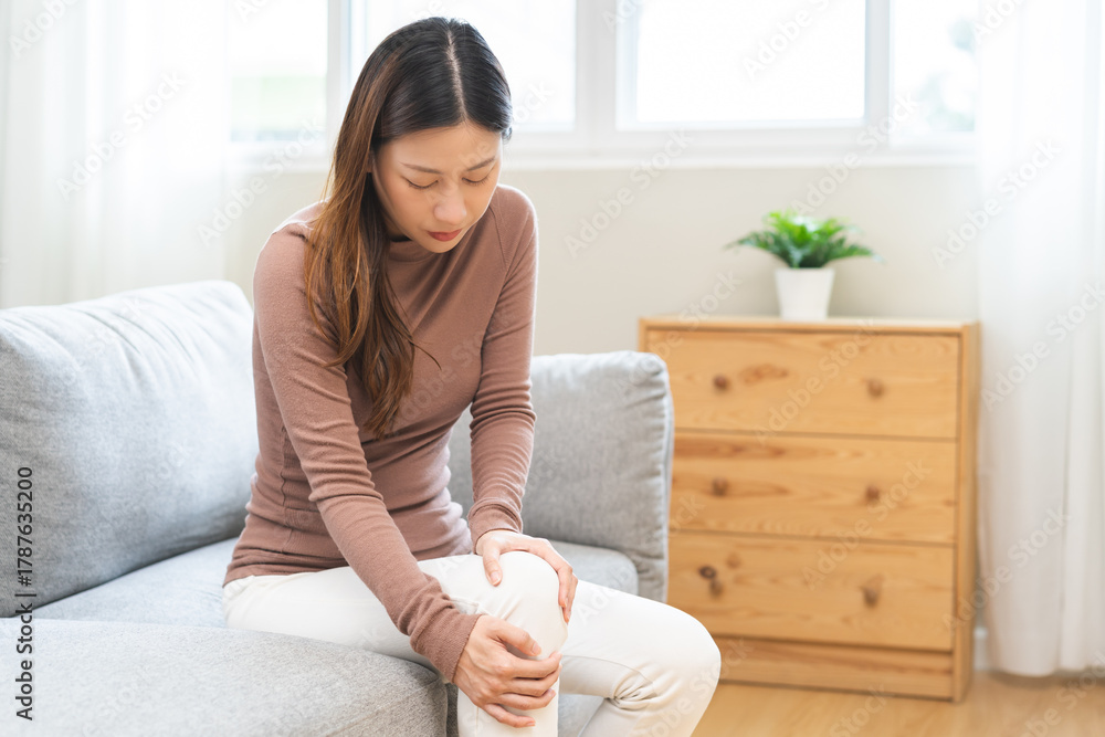 © KMPZZZ - Health care of knee pain concept, suffering asian young woman arthritis sitting joint ache, sore cramp or sprain tendon in leg on sofa at home. Osteoarthritis chronic disease of body problem, recovery