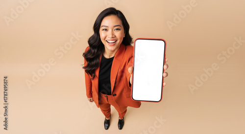 Smiling Asian woman holding a smartphone with a blank white screen for mockup, high angle view of a happy professional on a beige studio background.
