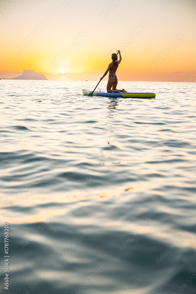 Naklejka premium Young woman paddleboarding in serene ocean at sunrise