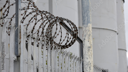 Industrial security scene featuring razor wire along a metal fence and weathered posts near large cylindrical silos, conveying vigilance, containment, and restricted access around an industrial site.