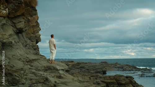 Young man standing on a rocky beach overlooking the sea with waves breaking on the shore and a stormy sky in the background.