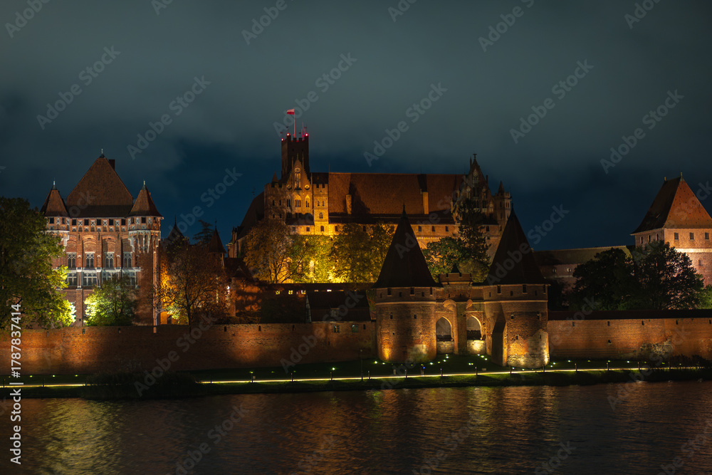 Obraz premium Nighttime Malbork Castle illuminated along the Nogat River, Poland