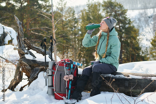 Caucasian young adult woman sitting on fallen tree trunk in snowy forest drinking from thermos, resting during winter hike with trekking poles and large backpack placed nearby