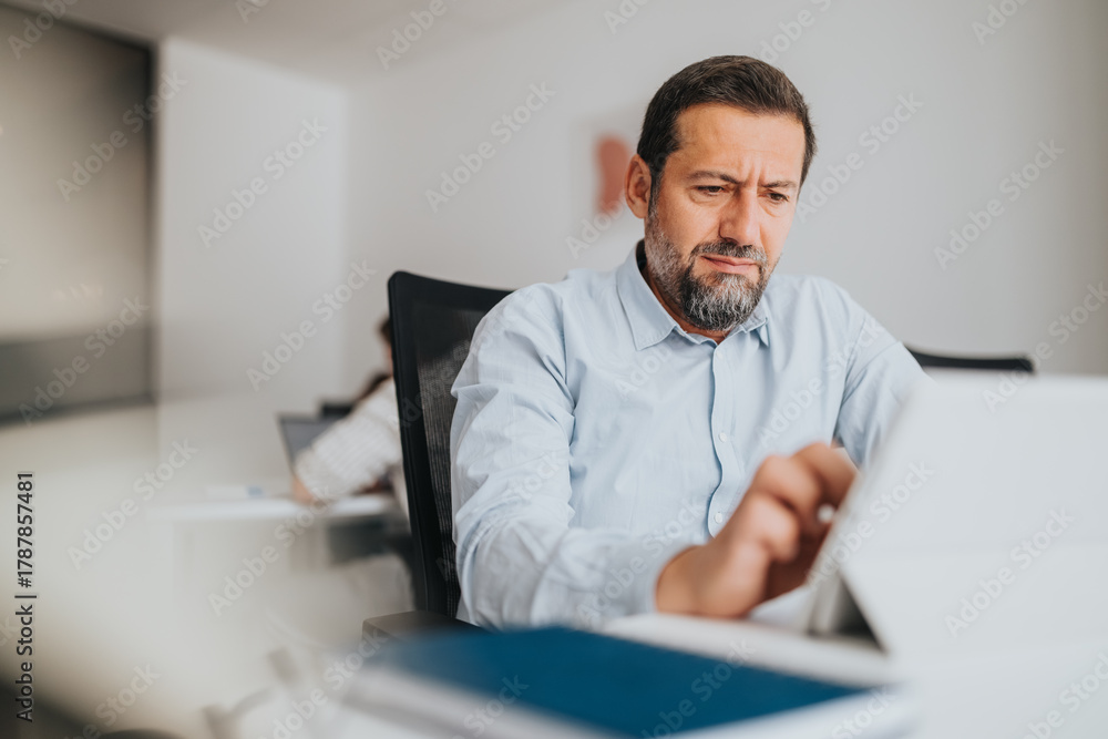Naklejka premium A professional man sits at a desk in a bright, modern office, concentrating on a tablet. The calm atmosphere and focused expression convey productivity, digital work, and everyday office routine.