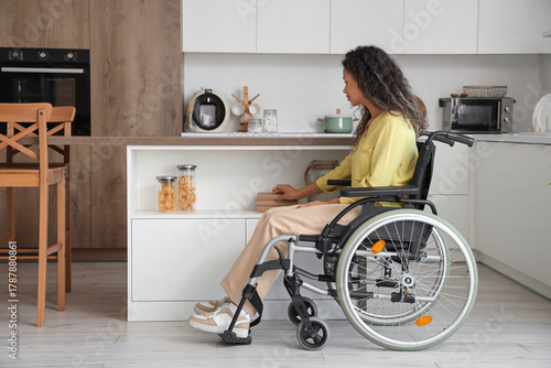 Young African-American woman in wheelchair taking book from shelf at home
